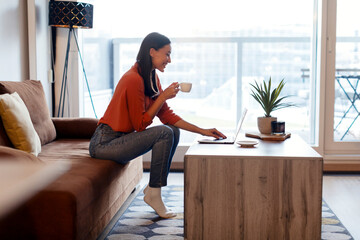 Profile side view of happy woman using laptop and drinking coffee, sitting at table, watching...