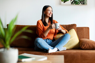 Fototapeta premium Happy European lady sitting on comfortable sofa, drinking coffee and looking away, thinking about plans, resting in modern cozy living room
