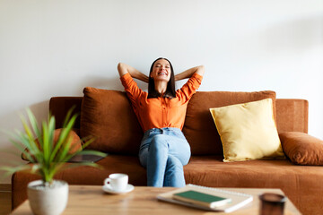 Relaxed peaceful lady sitting on cozy couch in living room, woman with hands behind head leaning...