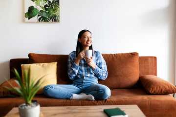 Attractive happy woman enjoying lazy morning with coffee, relaxing on couch with cup of hot drink at home, full length, free space