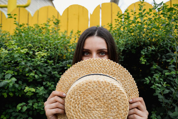 Young woman playfully hiding behind a straw hat amidst lush greenery in a vibrant garden setting