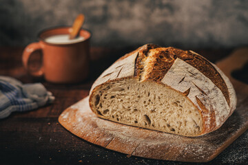 A rustic artisan sourdough bread loaf accompanied by a mug of milk and a striped linen napkin, creating a warm, inviting kitchen atmosphere. Suitable for baking, breakfast, or food styling themes
