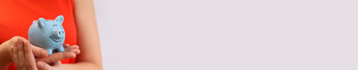 Piggy bank in female hands with copy space. Blue piggy bank, concept of saving money and financial planning. Woman in red t-shirt with piggy bank on light background. Cute blue piggy bank. Banner