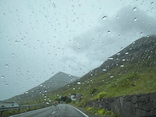 The windshield of a vehicle is covered in rain drops. Views beyond to a curved two-lane road...