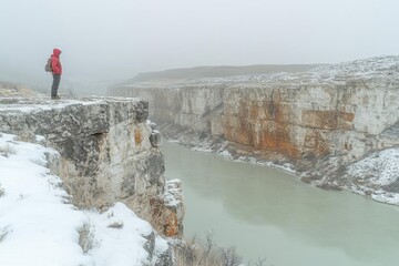 Hiker pauses on snowy cliff overlooking a misty river canyon during a winter storm