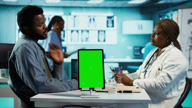Black medic meets with patient next to isolated mockup display at the office, doing routine consultation and reviewing records for a treatment plan. Physician at private clinic. Camera B.