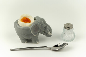 Das perfekt weich gekochte Ei in einem lustigen Eierbecher in Tierform (Elefant) mit Salzstreuer und Eierlöffel, weißer Hintergrund, für Freude und Spaß beim frühstücken, Platz für Text
