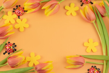 Obraz premium wunderbare Rot-gelbe Tulpen, Marienkäfer aus Schokolade und gelbe Blüten aus Filz bilden einen Rahmen, gelber bis oranger Hintergrund, mit Platz für Text in der Mitte