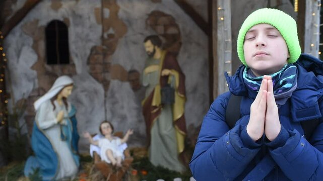boy in blue winter jacket praying in front of nativity scene with holy family figures outside.	