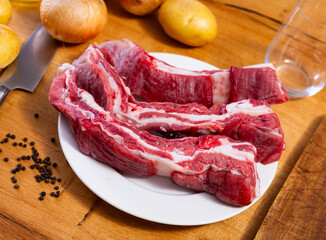 Raw beef ribs lie next to vegetables and spices - process of cooking homemade festive dish. Unpeeled vegetables, spices, olive oil and cooking utensils are placed on table before processing ingredient