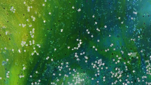 Green liquid with glitter and stars slowly flows down, mixing colors, abstract background. Green, blue shades.