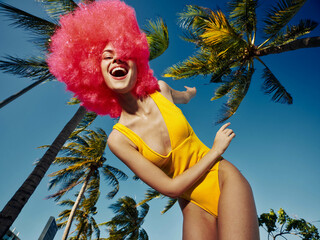 Bright pinkhaired woman in a yellow swimsuit poses in front of lush palm trees under a clear blue...