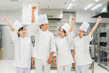 Obraz premium 厨房・キッチンに立つコックコート姿の調理師・シェフ・コックの男女（ガッツポーズ） 