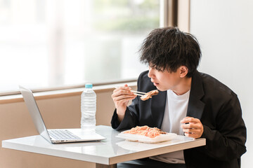 イートインスペース・社員食堂でパソコンを見ながら昼食にお弁当を食べる若いアジア人男性ビジネスマン
