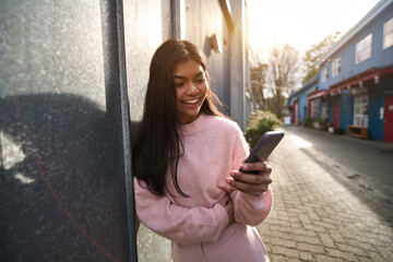 Candid photo of happy Asian teenage female model influencer using apps chatting on smartphone in the city. Smiling cool teen girl holding mobile cell phone looking at cellphone having fun outdoors.