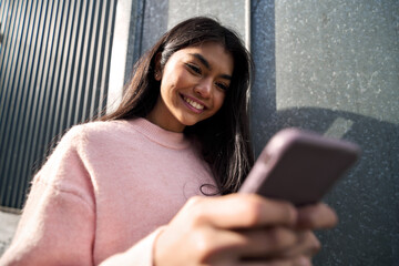 Candid photo of happy Asian teen female model influencer using apps typing chatting on smartphone in the city. Smiling cool girl holding mobile cell phone looking at cellphone having fun outdoor.