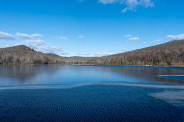 Serene winter lake scene. Partially frozen water reflects the clear blue sky and distant hills under a bright sun.