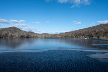 Serene winter landscape: a tranquil lake partially frozen, nestled amidst bare trees and hills under a clear blue sky.