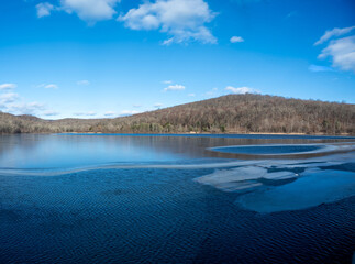 Serene winter lake scene. Partially frozen water, blue sky, and distant wooded hills create a...