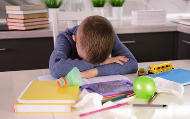 Frustrated, unhappy child sits down at table and does his homework among stack of books and...