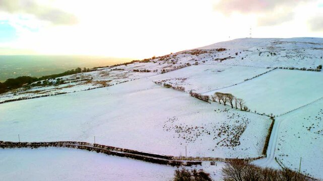 Aerial shot of Cavehill in Belfast, Northern Ireland on a snowy and calm winter day. The camera moves forward over the snowy mountain as the sun sets. Shot in 4K, 60fps and in Rec709 color.