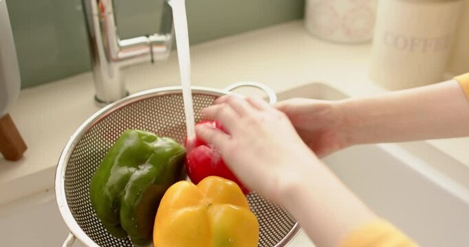 Washing colorful bell peppers in colander under running water in kitchen sink