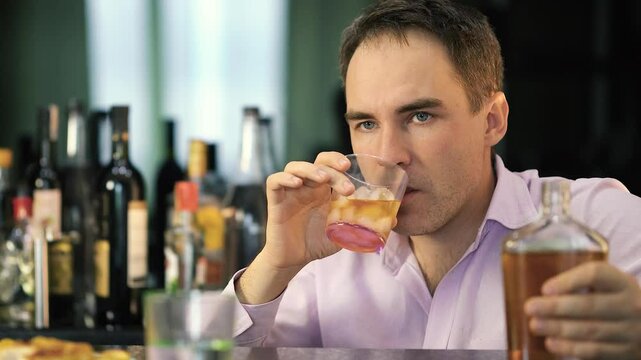 young man drinks whiskey from a glass in a bar on a dark background. A businessman with a bottle in his hands is relaxing in a pub with alcohol after work.