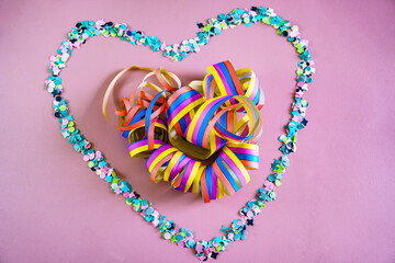 A heart made of colorful confetti frames streamers on a pink background. Carnival party concept. Flat lay, top view.