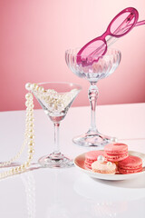 In front view, a dish of macarons, some glasses with a pearl string, sunglasses, and an empty space for product displays against a pink backdrop. A cosmetics advertisement with a party concept.