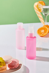 A front view of two pink bottles on a white surface. Surrounded by macarons, a cocktail glass filled with candy canes, and orange slices in glasses. The background is green. Commercial cosmetic image.
