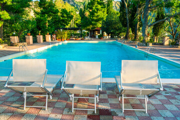 three white desk chairs or chaise lounge with blue swimming pool and hotel green garden on...