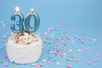 White coconut birthday cake on blue background with burning candles and many colorful sprinkles....