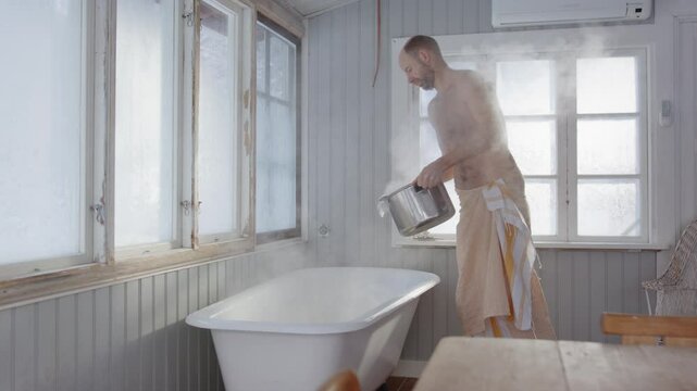 Bare chested man in towel pours boiling hot water from woodstove into steam bath