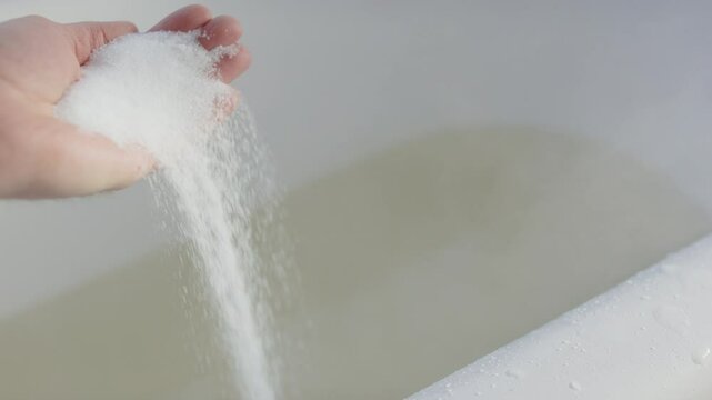 Epsom salts being dropped in slow motion from hand into steaming bath