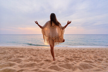 Concentrated calm young yoga woman with boho style with lotus posture hands on sandy beach enjoying...