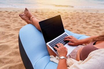 Black blank empty computer. Nomad girl freelancer lies on inflatable sofa on sandy beach at sunset...