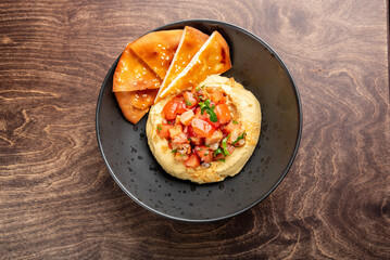 Delicious hummus topped with fresh diced tomatoes and herbs, served with crispy pita chips on a dark plate. Perfect for appetizers or healthy snacking.