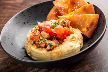 Delicious hummus topped with fresh diced tomatoes and herbs, served with crispy pita chips on a dark plate. Perfect for appetizers or healthy snacking.