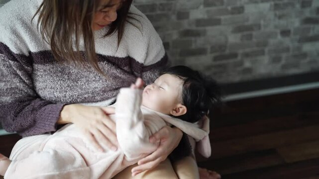 In a warm winter Japanese apartment room, a 6-month-old Japanese girl wearing fluffy clothes and her mother are sitting on the floor holding their sleeping baby.