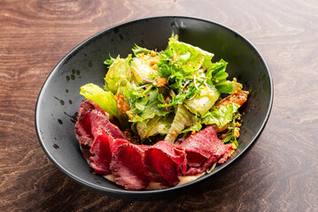 A close-up of a delicious salad featuring fresh greens and tender slices of roast beef in a dark bowl, set against a wooden background. Perfect for food-related content.