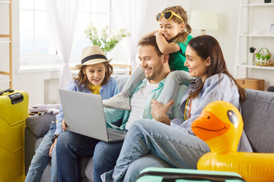 Young happy family booking tickets online via laptop in internet sitting on sofa in living room at home together. Parents with kids waiting for summer vacation trip. Travel concept.