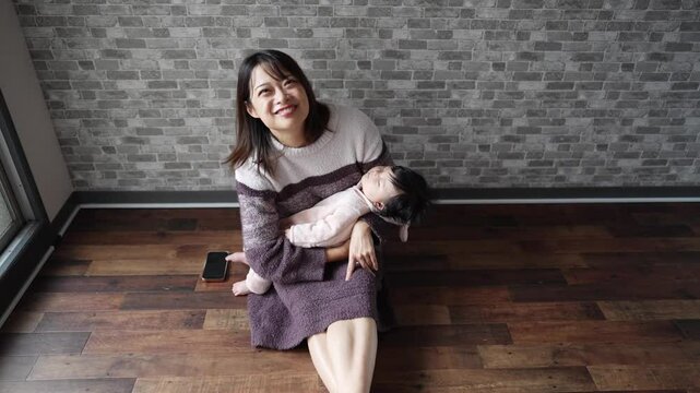 In a warm winter Japanese apartment room, a 6-month-old Japanese girl wearing fluffy clothes and her mother are sitting on the floor holding their sleeping baby.