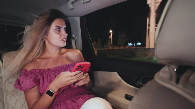 A young woman dressed elegantly in a stunning red dress is using her smartphone while comfortably seated in a luxurious car at night