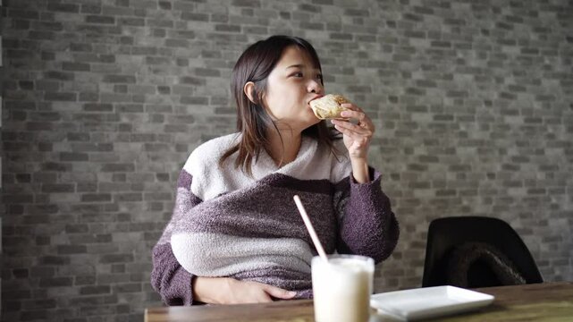 A six-month-old Japanese baby girl and her mother in fluffy clothes are sitting on a chair, breastfeeding and eating breakfast coffee and bread in a warm winter room. Slow motion video.