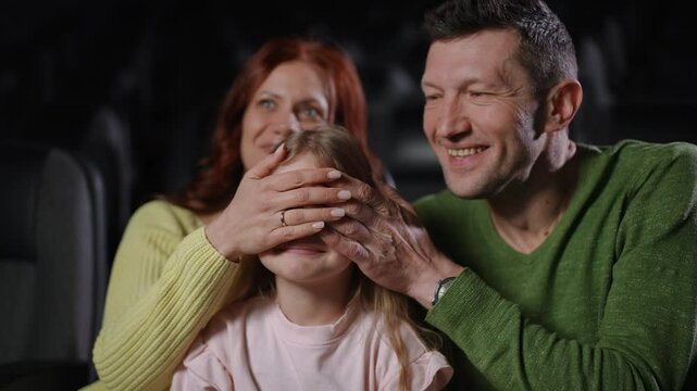 The family enjoys a playful moment in the cinema as the parents cover their little girls eyes, teasing her about the movie. The girl smiles, trying to push their hands away, while they share a
