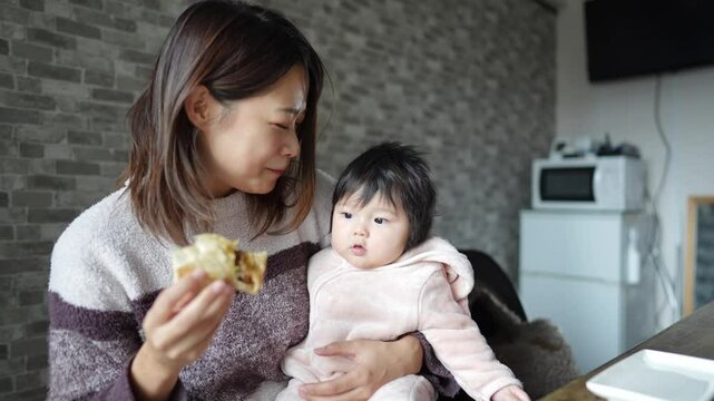 In a warm winter room, a six-month-old Japanese baby girl and her mother in fluffy clothes sit on chairs in an apartment room and eat a breakfast of bread and coffee. Slow motion video.