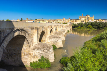 Fototapeta premium Roman bridge in Cordoba town in Spain. Ancient stone bridge spanning a river, with a historic city and cathedral in the background under a clear blue sky.