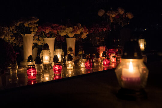 A Bunch Of Grave Candles Glowing With A Yellow Light On A Gravestone In The Background And One Grave Candle Glowing In The Foreground On All Saints' Day In The Night