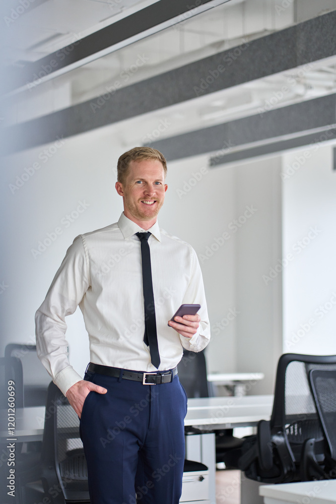 Poster Vertical portrait of smiling professional business man standing in office holding mobile cellphone. Young happy businessman entrepreneur using smartphone looking at camera with cell phone at work.