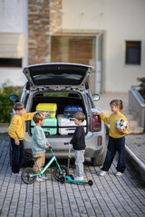 A happy family packs their car for a trip. Children help load luggage, a bicycle, and a scooter into the trunk, excited for their family vacation and outdoor adventure.